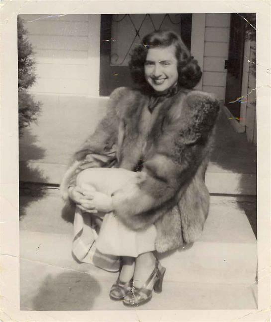 A woman dressed in a fur coat and high heels sits casually on the steps of a house, smiling.