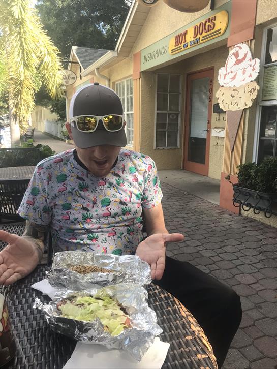 A man in casual attire sits at a cafe table, holding his hands up in surprise at his food.