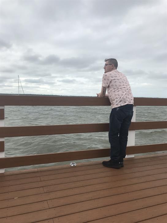 A man leans against the railing of a pier while looking out at the sea on an overcast day.