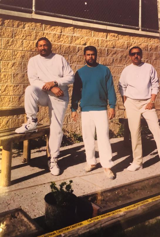 Three men stand together outdoors, enjoying a sunny day. The setting includes a textured stone wall.