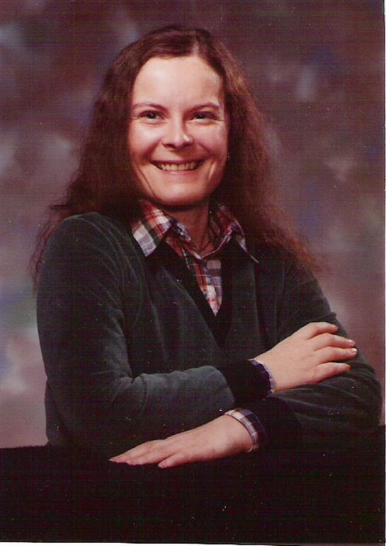 A woman with long hair smiles while resting her arms on a table.