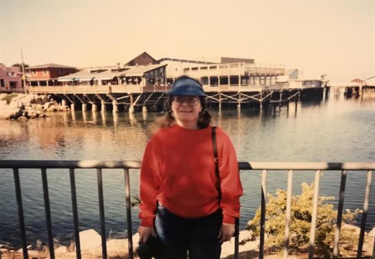 A woman in a red sweater and hat stands by a waterfront railing with a pier behind her.