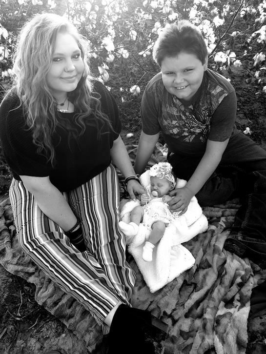 Two kids sit on a blanket, holding a baby in white on a sunny afternoon.