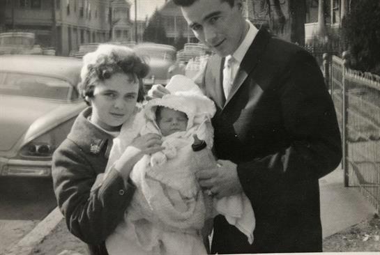 Couple cradles newborn baby wrapped in a blanket while standing on a lively street.