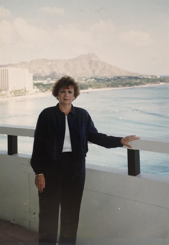 A woman poses on a balcony with a scenic view of the ocean, waves, and mountains during the day.