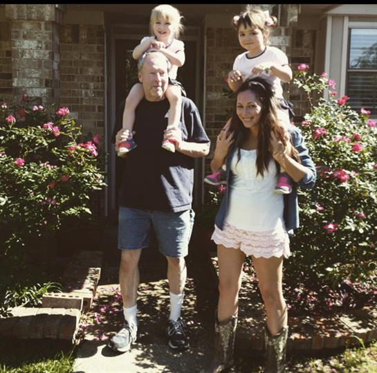 A happy family poses in a garden, with two kids on adults' shoulders, all smiling.
