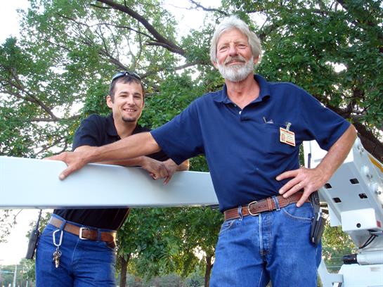 Two men stand outdoors, smiling and posing near a large panel in a green park area.