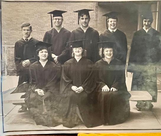 A group of eight graduates poses in traditional caps and gowns outside a brick building.