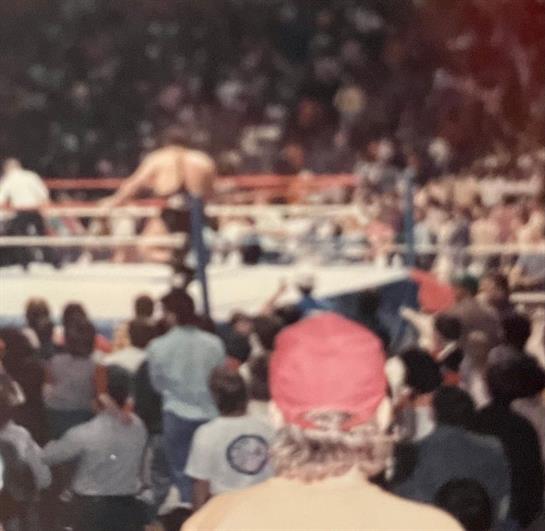Fans fill the arena, eagerly watching a wrestling match unfold in the ring as excitement builds.