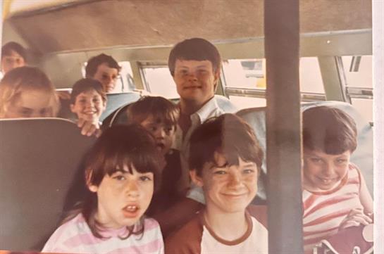A cheerful group of kids and adults share smiles on a bus ride in a retro setting.