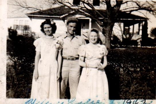 Three people stand together outside, smiling in vintage clothing, near a house in the background.