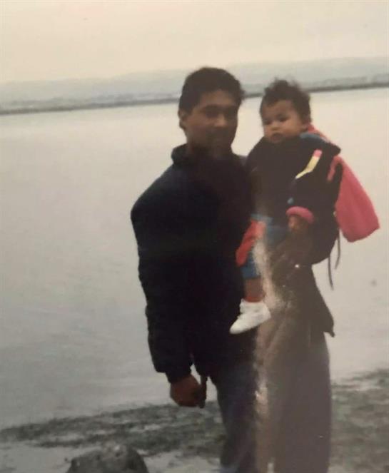 A father stands by the water holding his young child while showcasing a recent fish catch.
