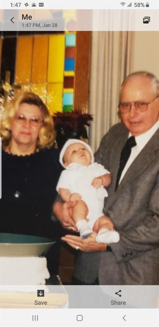 An elderly couple smiles while holding a baby dressed in white attire in a colorful setting.
