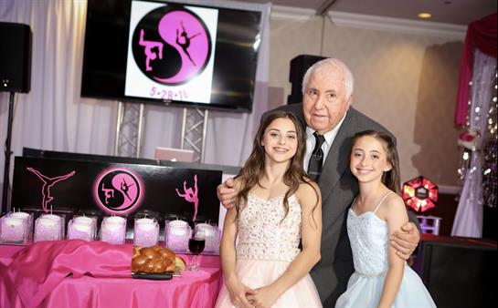 Two girls in formal dresses pose with an older man at a festive gathering with decorations.