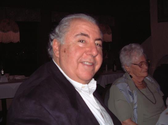 A joyful man is seated at a social gathering, smiling warmly with friends nearby in low light.