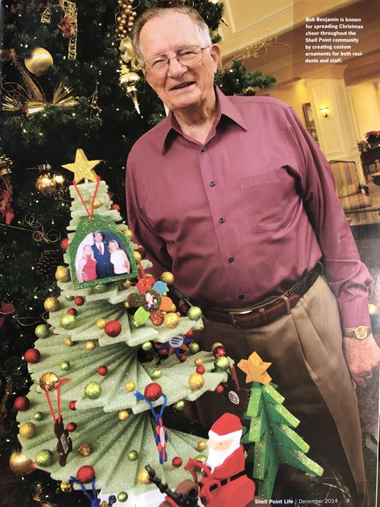 A senior man stands proudly next to a colorful Christmas tree adorned with decorations.