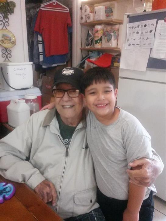 An elderly man sits smiling with a young boy, both looking happy and relaxed in a cozy room.