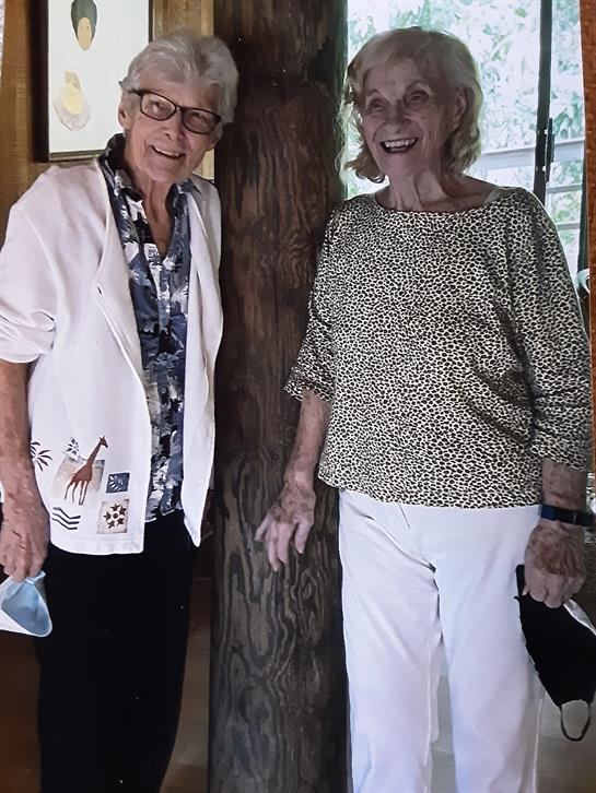 Two smiling women stand beside a tree indoors, enjoying each other's company in a cozy setting.