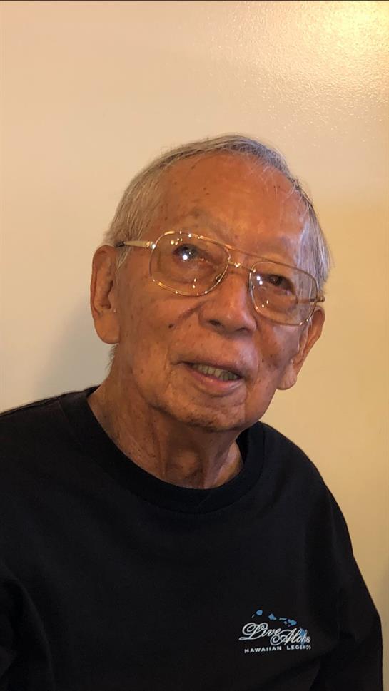 An elderly man wearing glasses sits comfortably, smiling and engaging in conversation indoors.