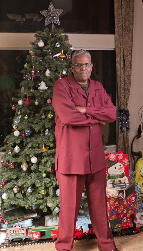 An elderly man poses proudly in burgundy pajamas beside a beautifully adorned Christmas tree.