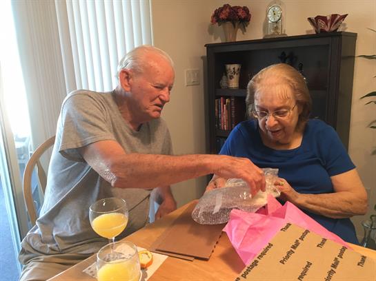 An elderly couple joyfully unwraps a surprise gift together at home, making happy memories.
