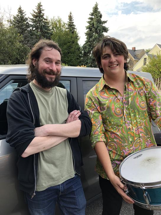 Two friends stand next to a car, one holds a drum, sharing smiles in a relaxed setting.