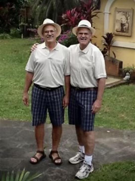 Two men stand side by side in a garden, both wearing matching outfits and smiling happily.