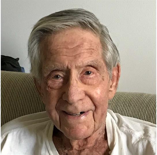 An elderly man with a warm smile relaxes on a couch in a cozy living room.