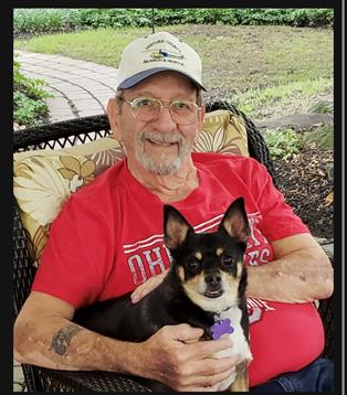 An elderly man smiles while holding his small black and white dog on a porch surrounded by greenery.
