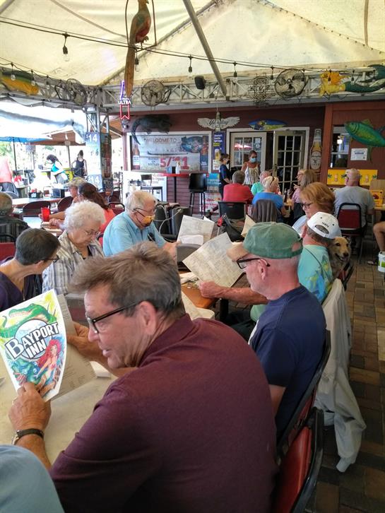 Groups of people gather around tables, looking at menus in a coastal restaurant setting.