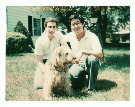 Two young men pose with a large dog in a green backyard on a summer day, enjoying the sun.