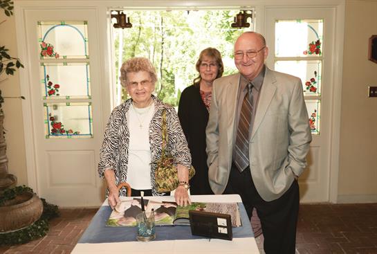 Three people interact around a scrapbook, sharing stories in a sunlit room filled with plants.