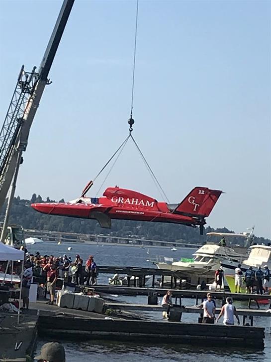 Bright red aircraft is being lifted from the water by a crane as spectators gather nearby.