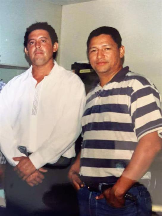 Two men stand closely side by side, dressed casually for a gathering in a relaxed environment.