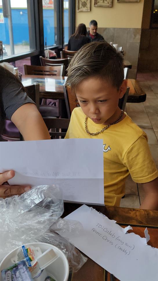 A young boy in a yellow shirt looks intently at an envelope held by an adult in a cafe.