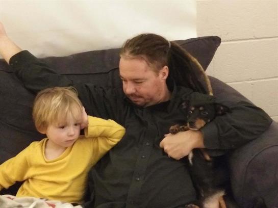 A man sits on a couch with a young boy and a small puppy, enjoying a warm family moment together.