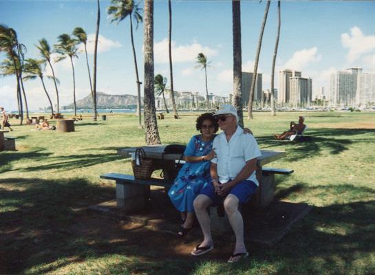 Elderly couple relaxes on a bench, enjoying their time together in a sunlit park near the beach.