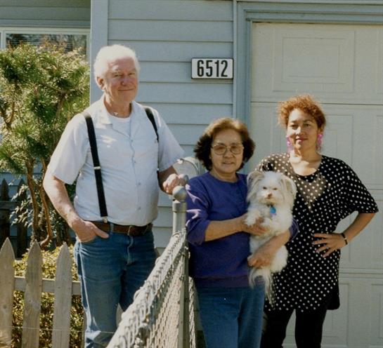 Three family members stand by the fence, holding a small dog, smiling in front of their house.