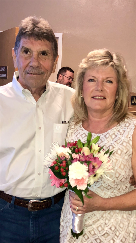 Smiling couple stands together indoors, surrounded by cheerful decorations and flowers.