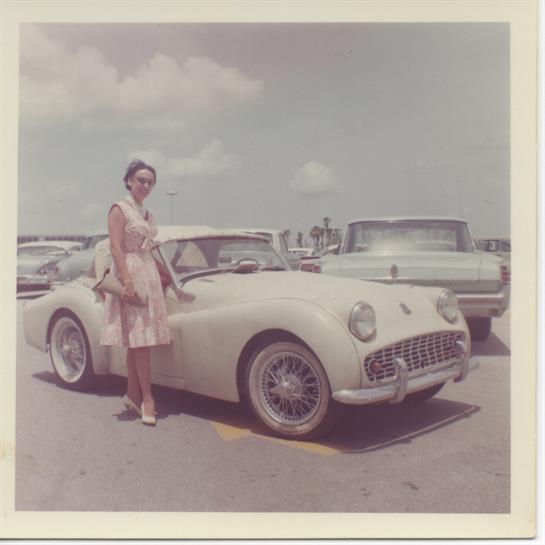 A woman in a pastel dress stands beside a classic convertible, surrounded by vintage cars.