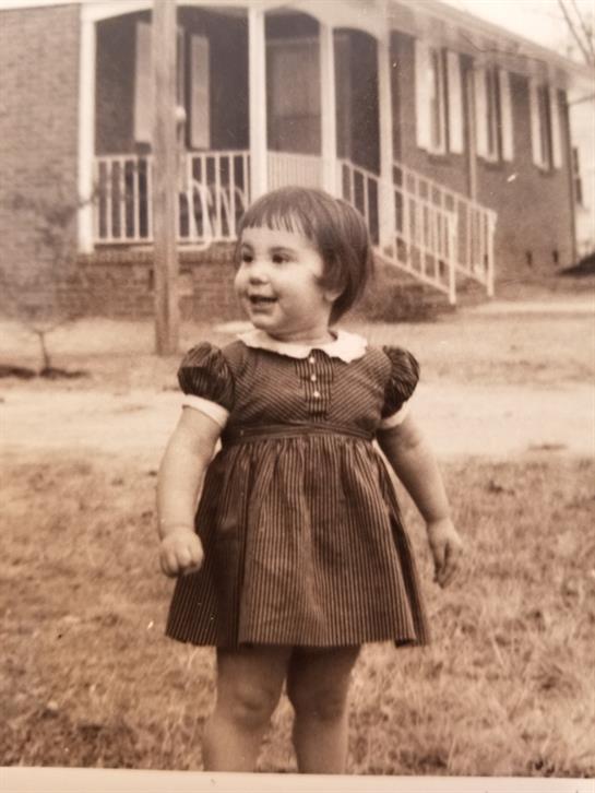 A cheerful young girl stands on grass in front of a house, smiling and enjoying the day.
