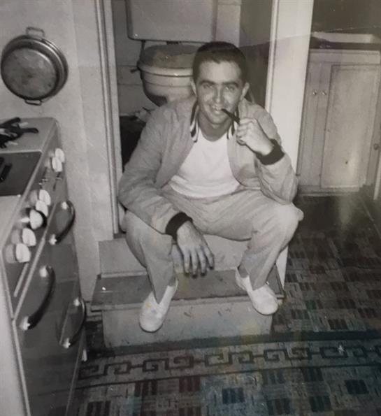 A man squats comfortably in a vintage kitchen, smoking a cigarette and looking relaxed.