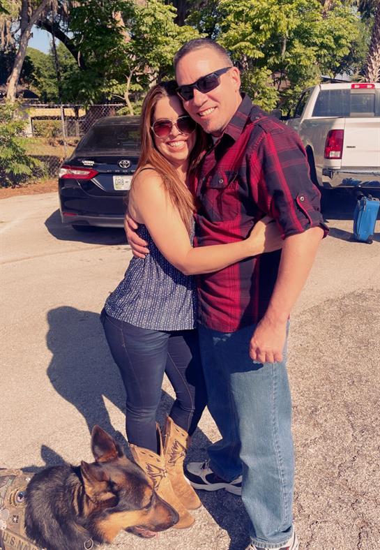 A joyful couple is hugging, dressed casually, enjoying a sunny day outdoors in a parking lot.