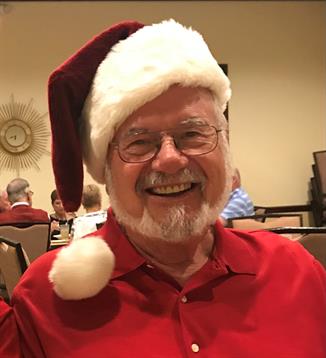 A cheerful man with glasses enjoys a holiday celebration at a community center, wearing a Santa hat.
