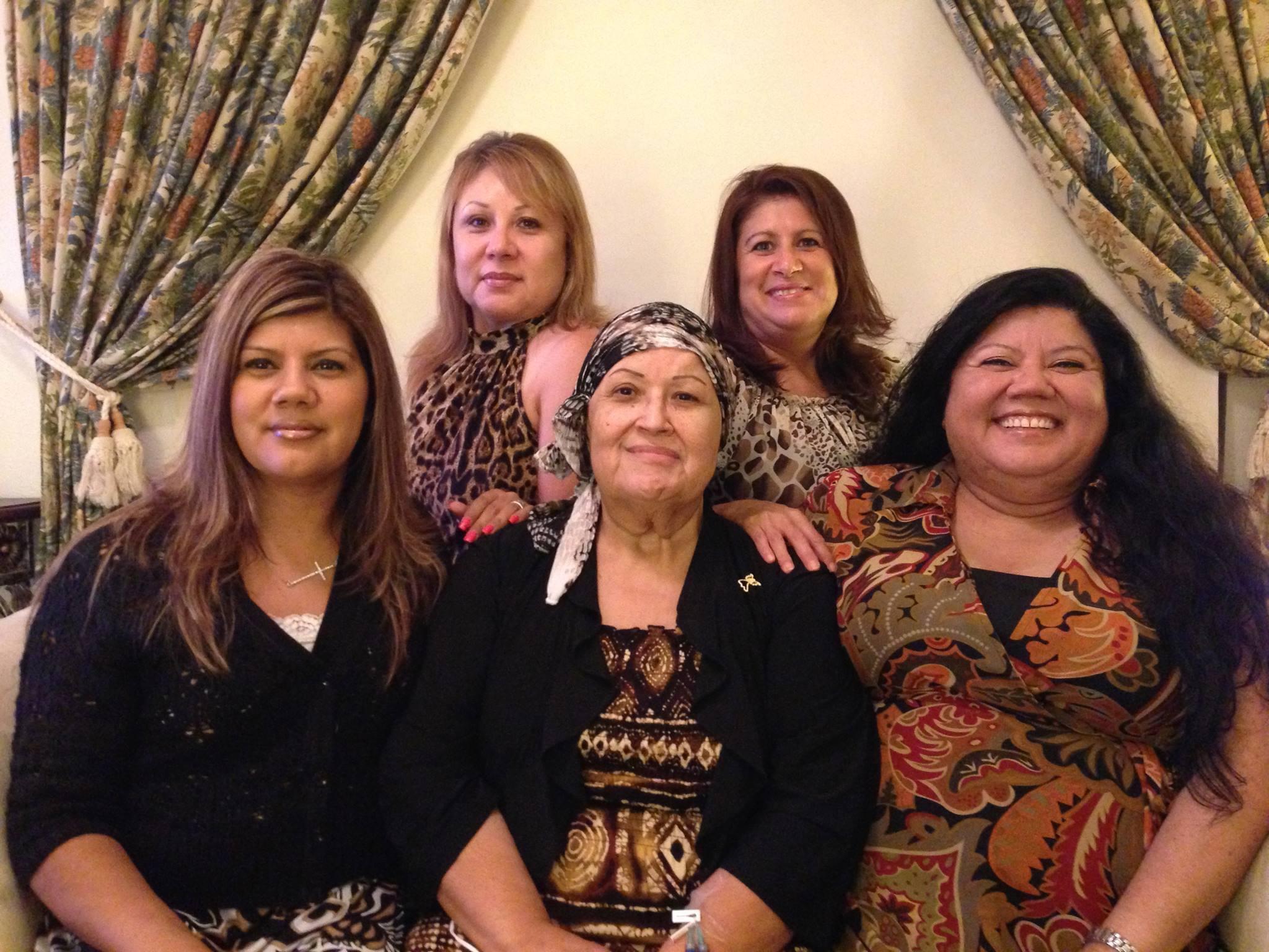 Six women pose together indoors, smiling and dressed in vibrant outfits, celebrating together.