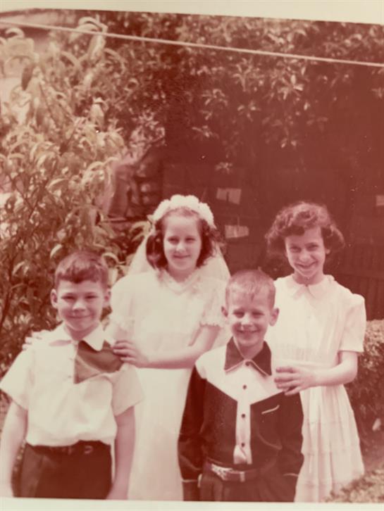 Four children stand together in a garden, dressed in vintage outfits, enjoying a sunny day.