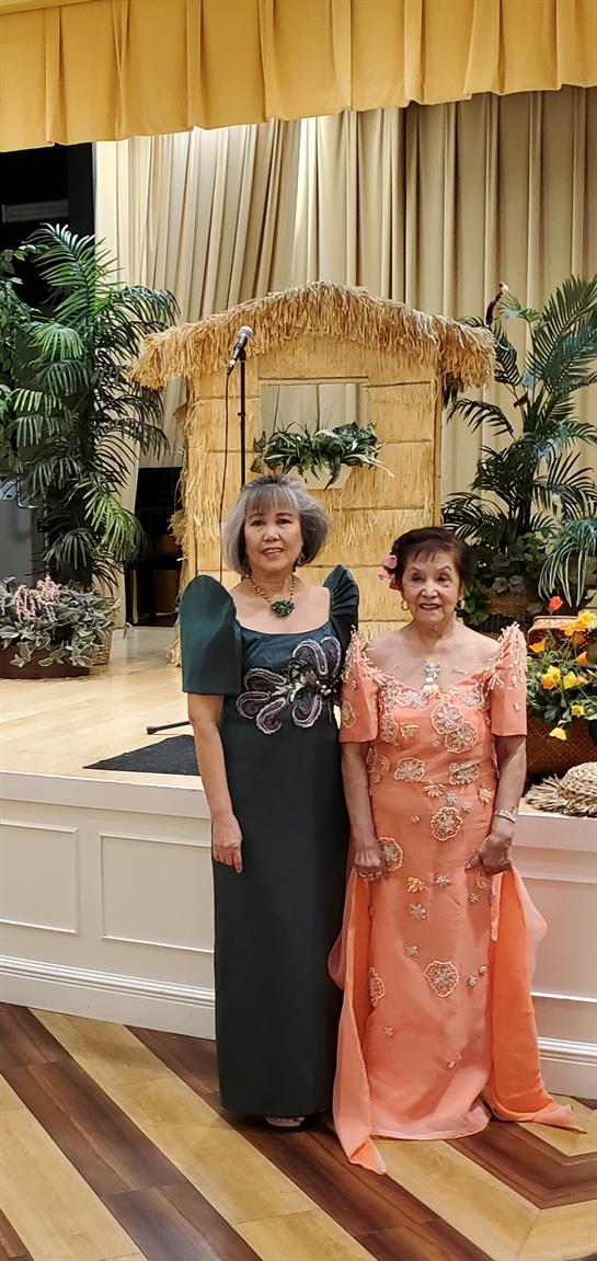 Women dressed in formal attire stand by a decorative tropical backdrop at an event.