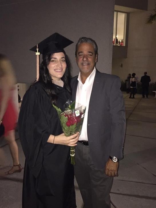 A graduate in cap and gown smiles with a family member, holding a bouquet.
