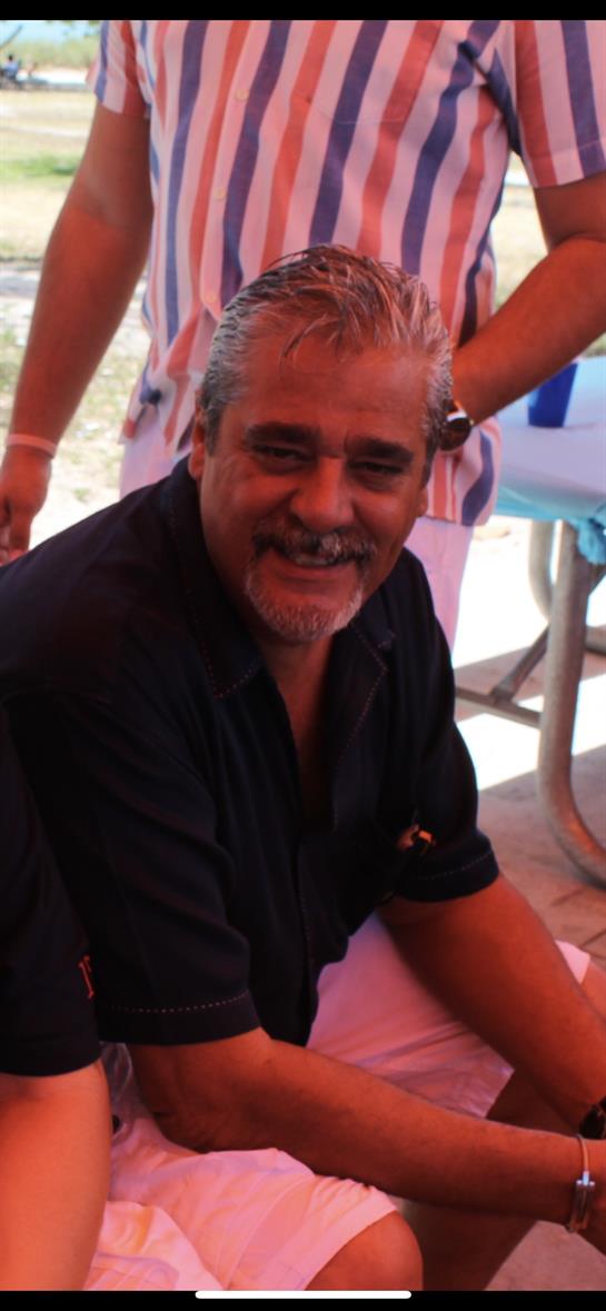 A cheerful man is seated, engaging with friends at an outdoor celebration in bright sunlight.