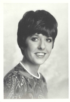 A smiling woman with short hair, pearls, and a patterned outfit in soft lighting.